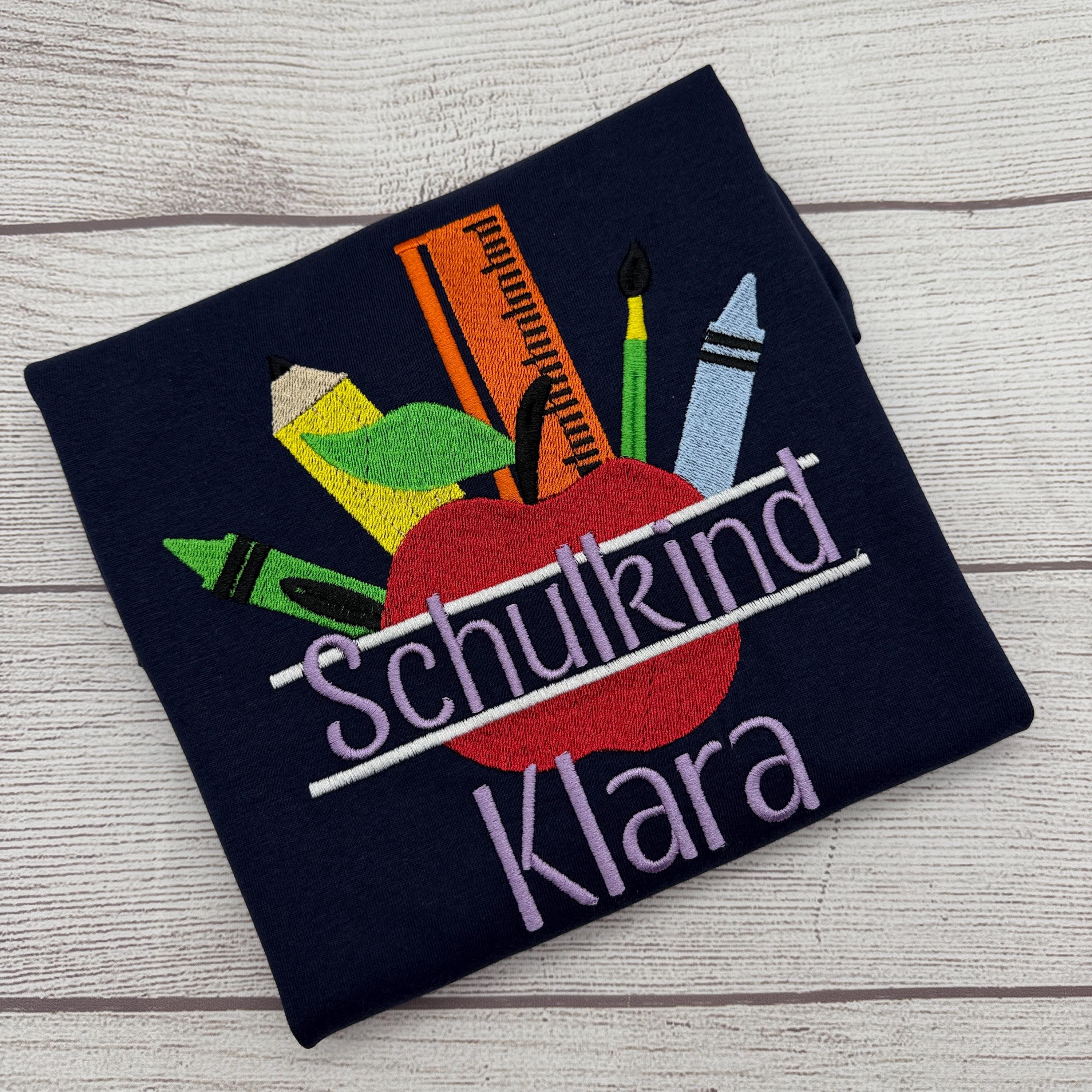 A dark blue t-shirt with a colorful design featuring a red apple, crayons, and a ruler, along with the text "Schulkind Klara" embroidered on it.