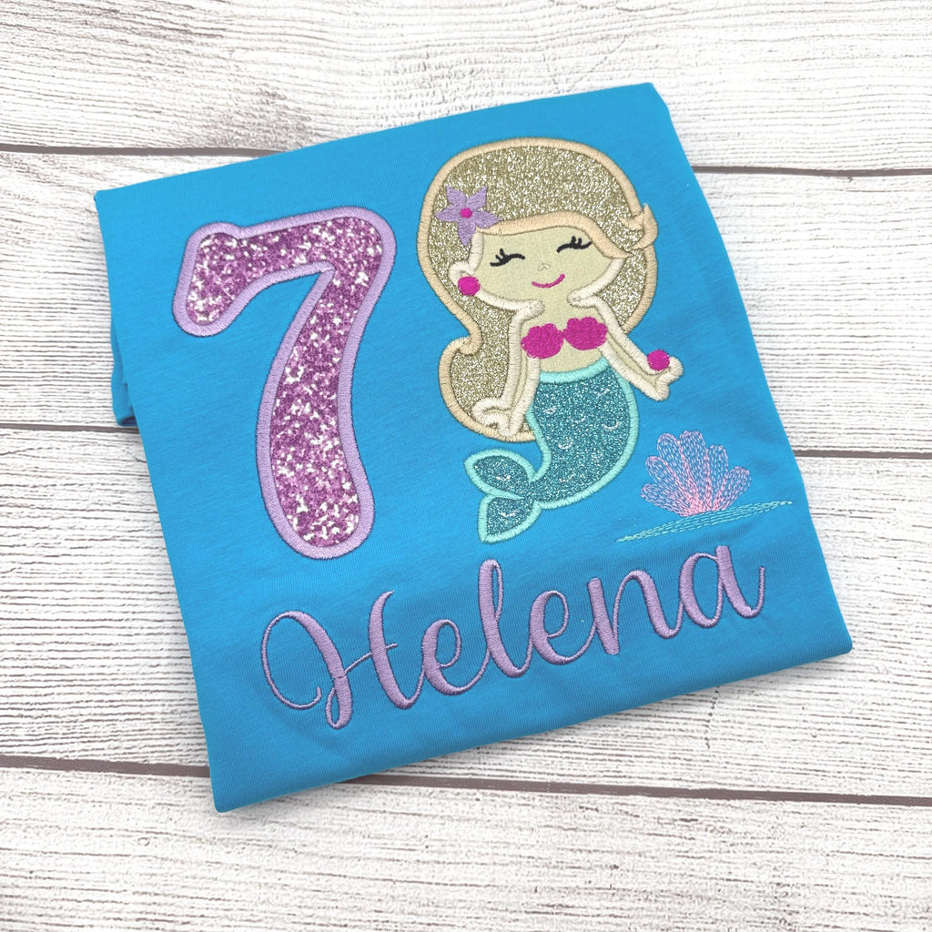 A blue t-shirt with a glittery number 7 and a mermaid design, along with the name "Helena" written on it.