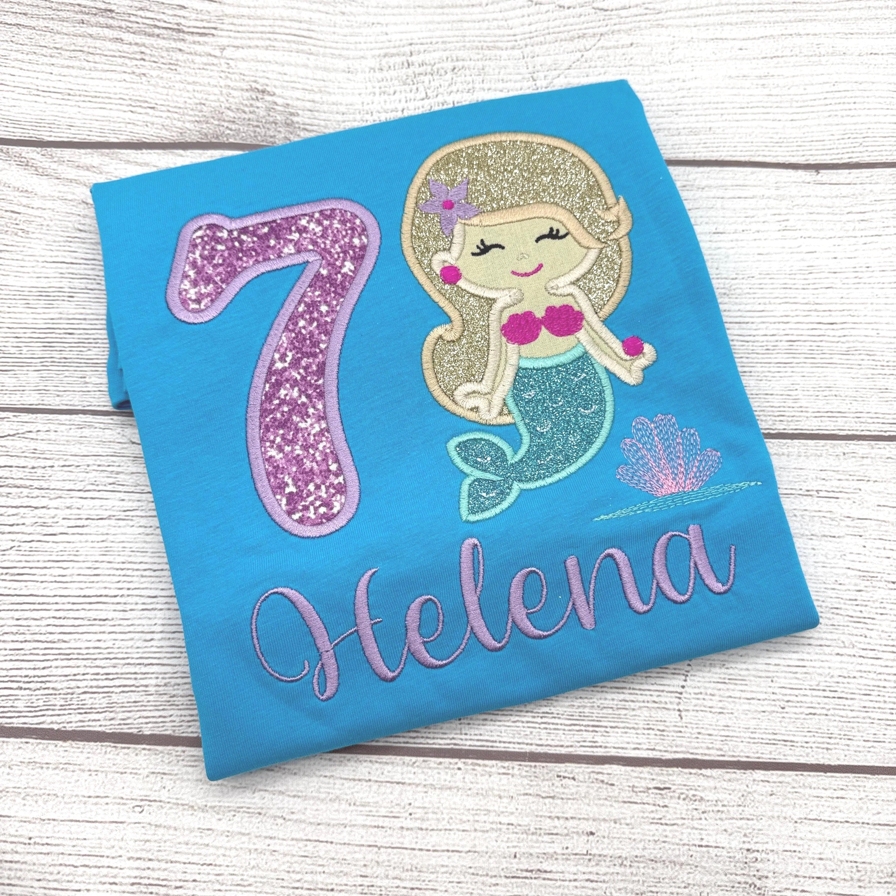 A blue t-shirt with a glittery number 7 and a mermaid design, along with the name "Helena" written on it.
